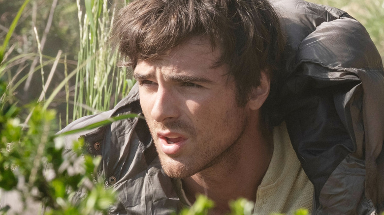 Jacob Elordi as Hig crouching behind vegetation in a grassy field in The Dog Stars