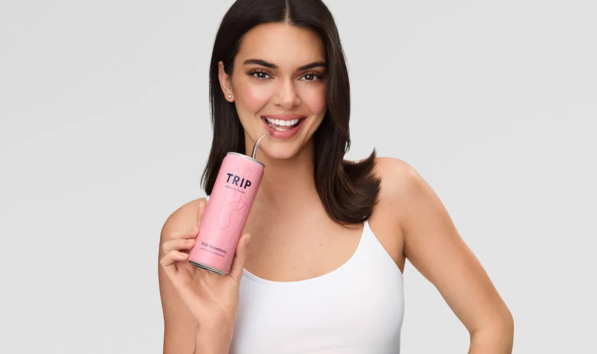 Kendall Jenner smiling while holding a pink "TRIP Wild Strawberry" CBD-infused drink with a straw.