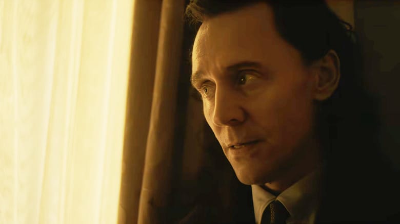Tom Hiddleston as Loki wearing a suit and standing by window drapes in Avengers: Doomsday