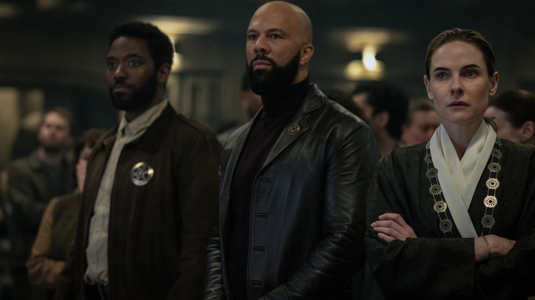 Chinaza Uche as Paul Billings standing in a crowded room next to Common as Robert Sims and Rebecca Ferguson as Juliette Nichols in Silo