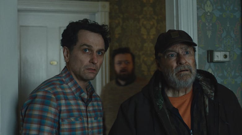 Matthew Rhys as Tom Loftis and Stephen Root as Wyck Crawford in Widow's Bay, standing next to each other with apprehension in a house, with Neil Casey as Kurt standing behind them