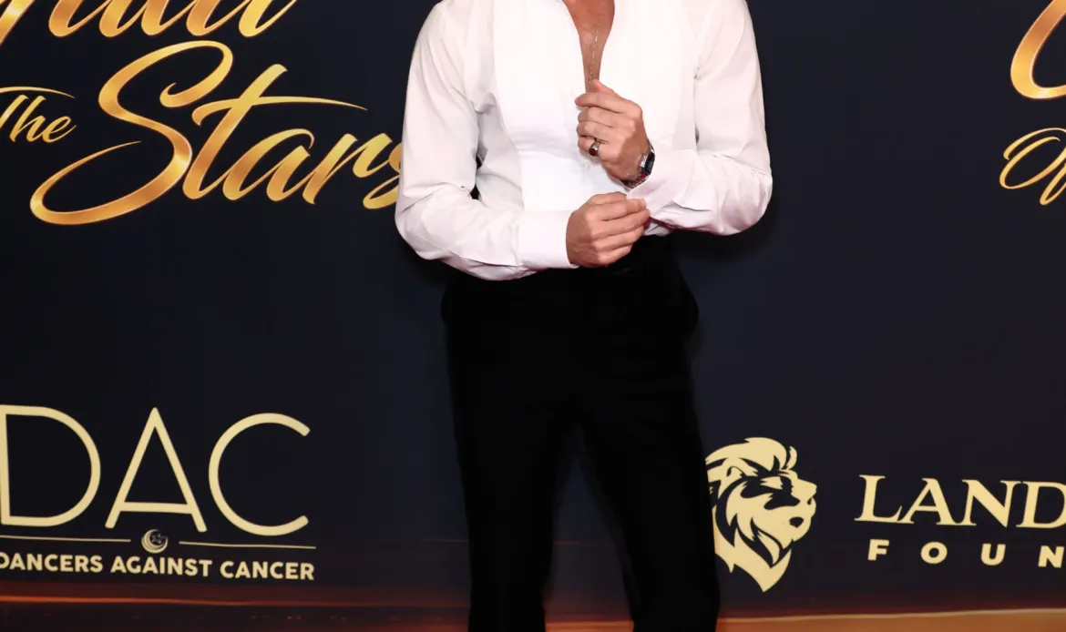 Sasha Farber poses on a red carpet in front of a "Gala of the Stars" step-and-repeat backdrop, wearing a white button-down shirt and black trousers.