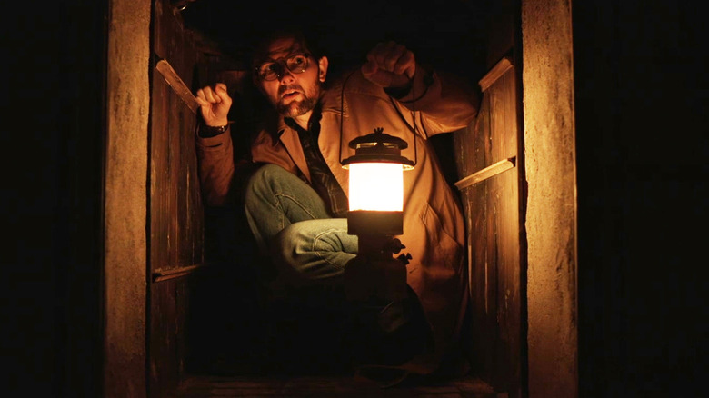 Adam Scott's Ohm Bauman is seen crouching in an enclosed space with a lantern in Hokum
