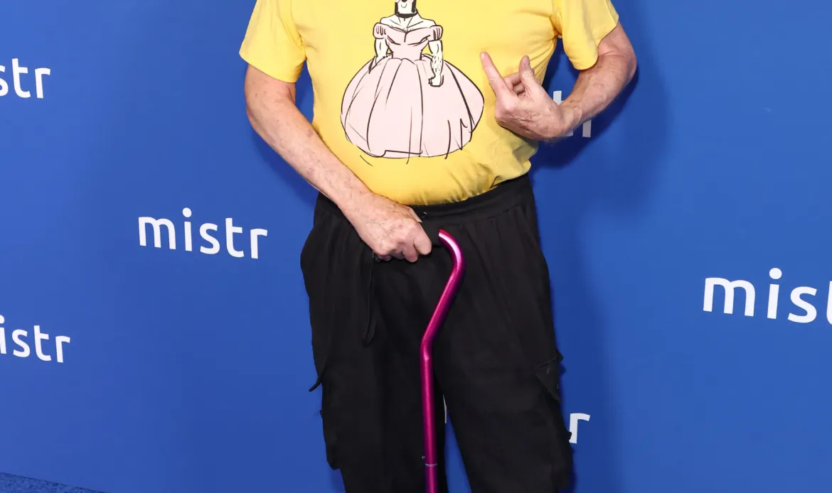 Bruce Vilanch poses on a blue carpet in front of a "mistr" branded backdrop, wearing a yellow graphic t-shirt, black pants, red clogs, and holding a purple cane.