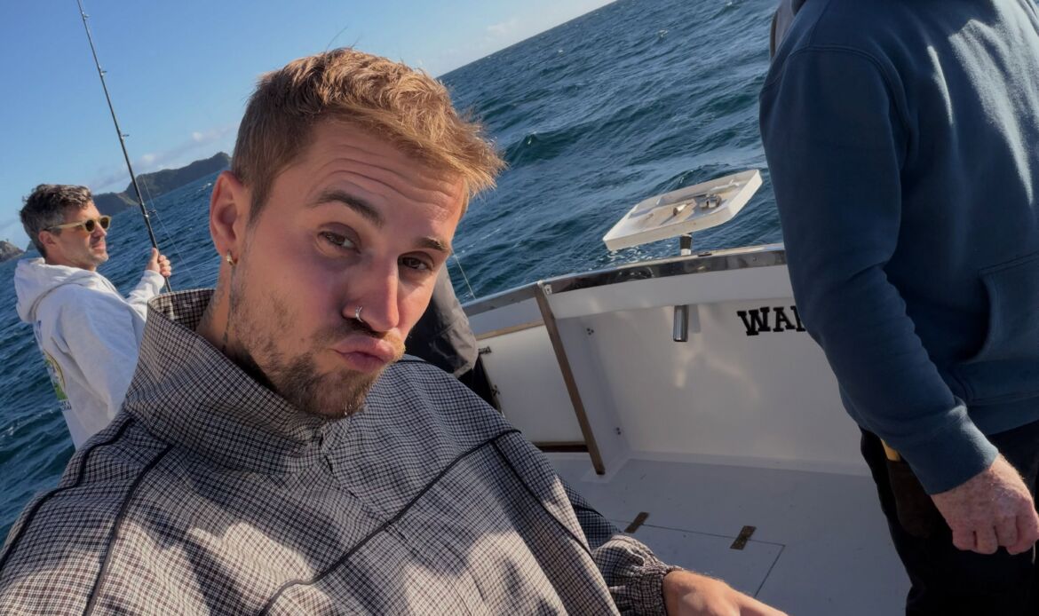 Justin Bieber takes a selfie on a boat while fishing, with two other men in the background.